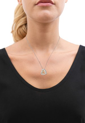 Collana con pendente a forma di cuore in argento, con un cuore interno dorato, indossata su una sottile catena, sopra una maglietta nera a V. Texture metallica liscia.