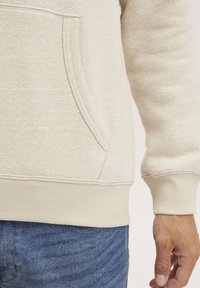 Beige genser med teksturert overflate, kengurulomme og ribbede mansjetter. Har et enkelt design og kontrasterende sømmer.