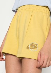 Žluté bavlněné šortky s elastickým pasem, s potiskem zlatého loga Nike a kulatým designovým detailem na nohavici.