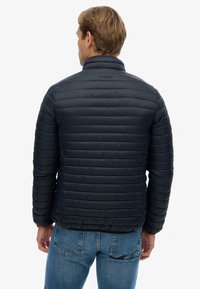 Navy puffer jacket with a quilted design, lightweight material, and a high collar. Features a subtle logo on the back, paired with denim jeans.