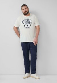 Witte katoenen t-shirt met blauwe grafische print, gecombineerd met marineblauwe broek en witte sneakers. Over het algemeen een ontspannen pasvorm en casual stijl.