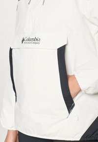 Bela lahka jakna s črnimi detajli, ki ima sprednji žep, elastične manšete in kapuco. Columbia logotip prikazan na prsih.