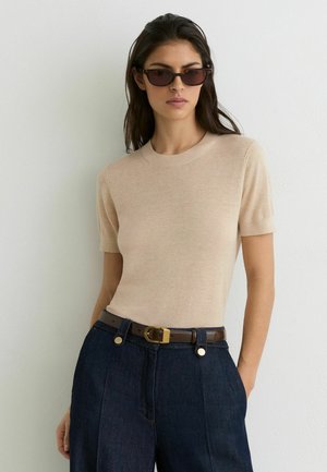 Kvinde iført beige kortærmet striktop, mørke bukser med højt talje og brun bælte samt mørke rektangulære solbriller, stående mod en hvid væg.