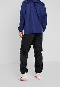 Veste imperméable bleu marine avec détails blancs, associée à un pantalon noir ample et des baskets blanches avec des accents rouges.