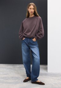 Mørk mauve sweatshirt med raglanærmer, kombineret med lyseblå, løstsiddende denimjeans. Modellen står med hænderne i lommerne og har brune flade sko på.