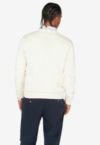 Suéter de punto color crema con dobladillo y puños acanalados, usado sobre una camisa de cuello azul claro. Debajo se pueden ver pantalones de un azul marino oscuro.