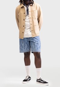 Chaqueta de lona beige claro con un bolsillo en el pecho, camiseta blanca, pantalones cortos de mezclilla azul con un patrón de llamas, zapatillas negras y calcetines blancos.