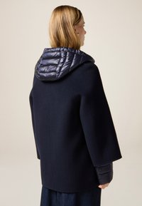 Cappotto blu navy con cappuccio imbottito, caratterizzato da una finitura testurizzata, vestibilità ampia e maniche larghe, con tessuto liscio in contrasto sul cappuccio.