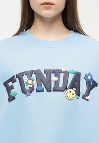 Μπλε φούτερ με μεγάλα μαύρα γράμματα που σχηματίζουν τη λέξη "FUNDAY", διακοσμημένο με πολύχρωμα χαμογελαστά λουλούδια, σύννεφα, αστέρια και καρδιές, σε ένα άτομο.