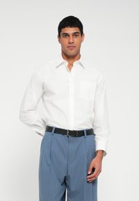 Weißes langärmeliges Hemd mit Brusttasche, kombiniert mit hellblauen, fein gestreiften Hosen und einem schwarzen Gürtel, vor einfachem Hintergrund.