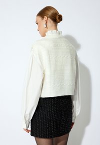 Weißer, kurz geschnittener Pullover mit strukturiertem Muster, über einer langärmeligen weißen Bluse getragen. Kombiniert mit einem schwarzen Rock mit Nadelstreifen.