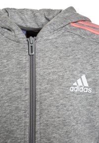 Felpa grigia con zip realizzata in un tessuto strutturato, con strisce rosa sulle spalle e un logo Adidas bianco sul petto.
