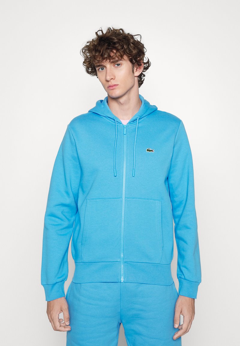 Sudadera con cremallera de color azul claro, hecha de una tela suave. Cuenta con capucha, bolsillos frontales y un pequeño logo en el pecho. Ajuste cómodo.