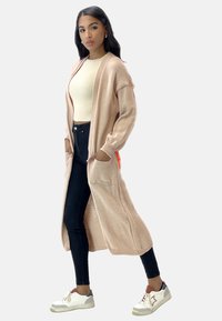 Lichtroze lange cardigan met geribbelde details, zijzakken, gedragen over een crème top en donkere jeans, gecombineerd met witte sneakers.