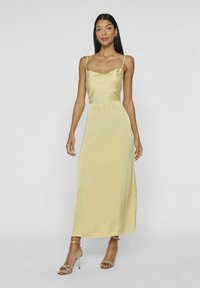 Vestido maxi de satén amarillo con escote en cowl, tirantes finos y una abertura lateral. Llevado con sandalias de tacón alto de tiras doradas.
