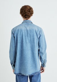 Camicia in denim azzurro chiaro con maniche lunghe, colletto a punta e polsini abbottonati. Presenta una vestibilità rilassata e un orlo leggermente curvato.