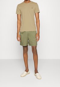 Vaaleanruskea puuvillainen t-paita, jossa on pieni logo, yhdistettynä oliivinvihreisiin pellavashortseihin. Mallilla on päällään beigeet liukuväriset kengät. Yksinkertainen, rento muotoilu.