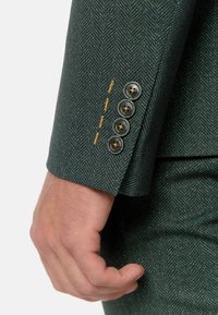 Ärmel eines grün gemusterten Sakkos mit fünf dunklen Knöpfen und kontrastierenden gelben Nähten an der Manschette. Glatte Stoffoberfläche.