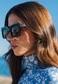 Zwarte oversized zonnebril met vierkante monturen, reflecterende lenzen en een logo aan de zijkant; gedragen met een blauwe bloempatronen top met hoge hals.