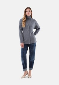 Grau gestrickte Strickjacke mit hohem Kragen, Knöpfen vorne und langen Ärmeln, kombiniert mit dunkelblauen Jeans mit aufgerolltem Saum und hellen Absätzen.