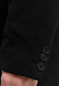 Mustvärviline villane jopevarrukas, millel on pehme tekstuur ja kolm musta nööpi, millel on igaühel neli auku, paigutatud cuffi lähedale.