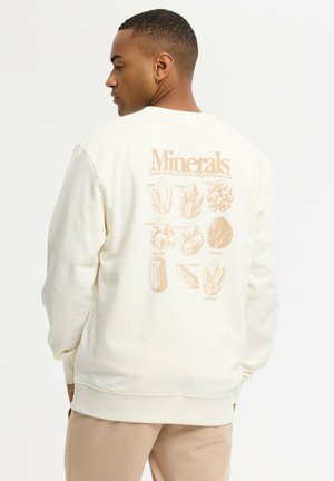Hombre de perfil con una sudadera blanca con ilustraciones y nombres de minerales en beige impresos en la espalda.