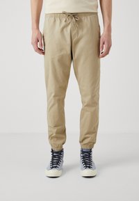 Beige, lichte joggingsbroeken met elastische cuff en verstelbaar trekkoord in de taille, gecombineerd met blauw-grijze high-top sneakers.