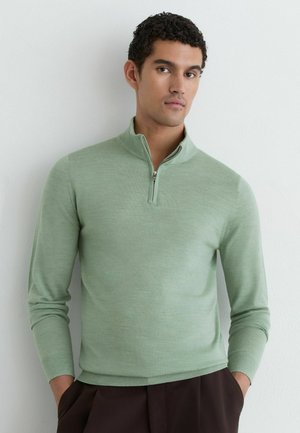 Mand iført en lysegrøn quarter-zip sweater og mørke bukser, stående med hænderne i lommerne mod en ensfarvet hvid væg.