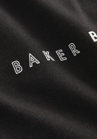 Čierna látka so štruktúrovaným povrchom, na ktorom je biele vyšité písmo s nápisom "BAKER." Detailný záber zvýrazňuje materiálové detaily.