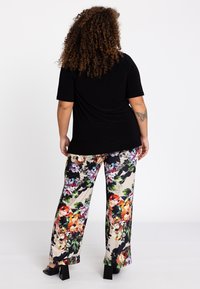 Zwarte top met korte mouwen, gecombineerd met bloemenmotief, wijde broek in levendige kleuren op een lichte achtergrond. De broek heeft een gladde textuur en opvallend patroon.