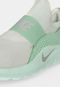 Близък план на бяла и мента зелена обувка Nike без връзки с текстуриран нос, перфорирана страна и широк ластичен презрамка с логото на Nike отгоре.