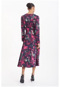 Dlouhý rukáv, květinové šaty v tmavě navy barvě s růžovými a zelenými vzory, s přiléhavým pasem a splývavou sukní, doplněné černými podpatky.