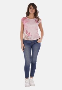 T-shirt rosa a maniche corte con motivi floreali in rosa e oro, abbinato a jeans blu aderenti e sneakers bianche. Tessuto misto cotone.