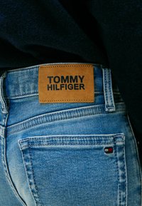 Vaqueros de mezclilla en azul claro con una etiqueta de cuero que presenta "TOMMY HILFIGER" en relieve en negro. Incluye un pequeño logo en rojo, blanco y azul.