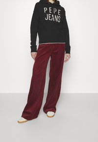 Sweat à capuche noir avec le texte "PEPE JEANS" en blanc, associé à un pantalon large en velours côtelé bordeaux et des chaussures plates de couleur claire.
