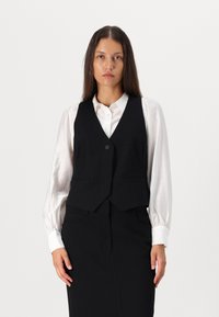 Black fitted vest with two front pockets, single button closure, worn over a white, long-sleeve blouse. Smooth fabric texture.