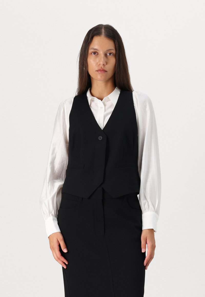 Black fitted vest with two front pockets, single button closure, worn over a white, long-sleeve blouse. Smooth fabric texture.