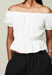 Haut blanc à épaules dénudées avec une finition texturée, col élastique froncé et ourlet peplum ; associé à un pantalon noir ample.