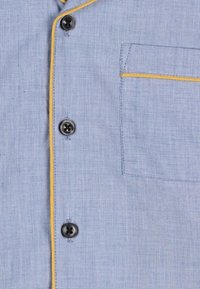 Chemise à motif bleu clair avec des boutons noirs, passepoil jaune le long des bords et une poche poitrine. La texture semble tissée et douce.