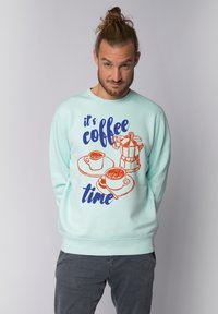 Sweat-shirt vert menthe avec des graphismes sur le thème du café en orange et marine, portant le texte "c'est l'heure du café" et des illustrations de tasses et d'une machine à café.
