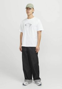 Hvid t-shirt med grafisk print, løs siddende mørke jeans, grå opfoldede ærmer og lysegrøn kasket. Stylings med sølvfarvede træningssko.