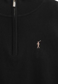 Svart stickad tröja med hög krage och dragkedja. Har en liten beige broderad golfspelare-logga på vänster bröst.