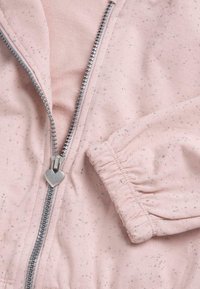 Tissu rose avec des paillettes argentées, montrant une fermeture éclair argentée partiellement ouverte avec un tirette en forme de cœur et un poignet élastique froncé.