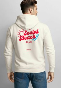 Cremefarbener Kapuzenpullover mit dem Schriftzug "Social Beach Club" in roter und blauer Schrift sowie Grafiken von Sonne und Welle. Weicher Stoff mit Kängurutasche, kombiniert mit blauen Jeans.