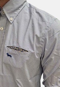 Camicia azzurro chiaro con piccoli pois blu scuro, colletto button-down e taschino sul petto con un rivestimento a contrasto e un logo blu navy.