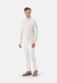 Cremfarbener Grobstrickpullover mit hohem Kragen, kombiniert mit weißen Hosen und beigen Stiefeletten. Die Textur ist dick und strukturiert.