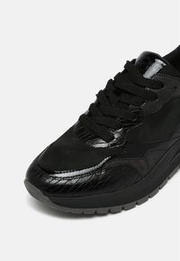 Baskets noires en cuir texturé, panneaux en daim, lacets noirs épais et semelle en caoutchouc grise sur fond blanc.