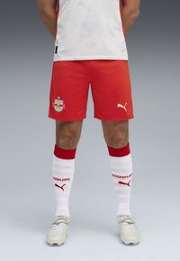 Pantalones cortos deportivos rojos en un vibrante rojo con un logo; combinados con calcetines blancos que tienen acentos rojos y texto, y botas de colores claros.