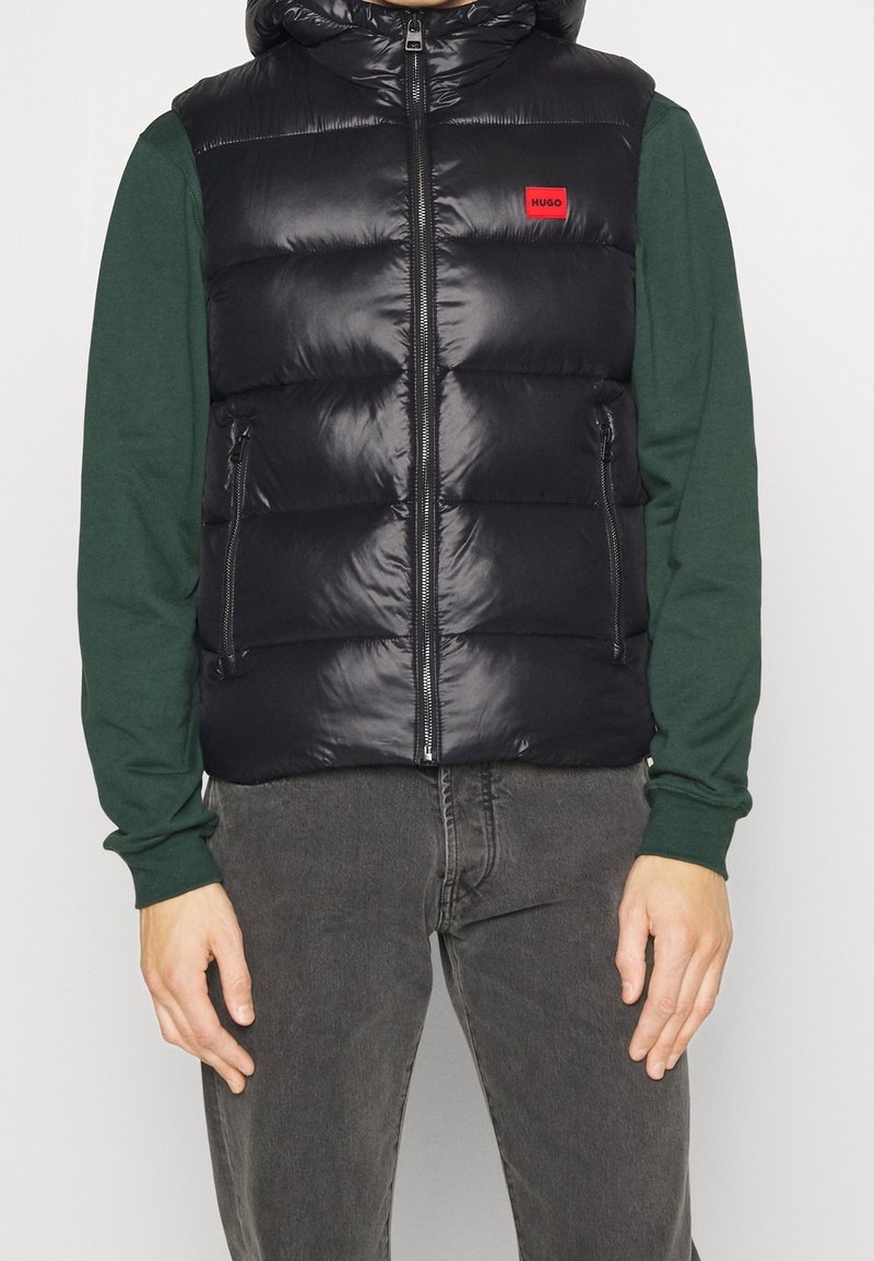 Schwarze gesteppte Weste mit glänzendem Finish, ausgestattet mit einem Frontreißverschluss und seitlichen Taschen, getragen über einem grünen Langarmshirt. Rotes Logodetail sichtbar.