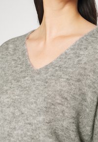 Grå, mjukstickad tröja med V-hals. Har en något fuzzig textur och en avslappnad passform som visar subtila färgvariationer.